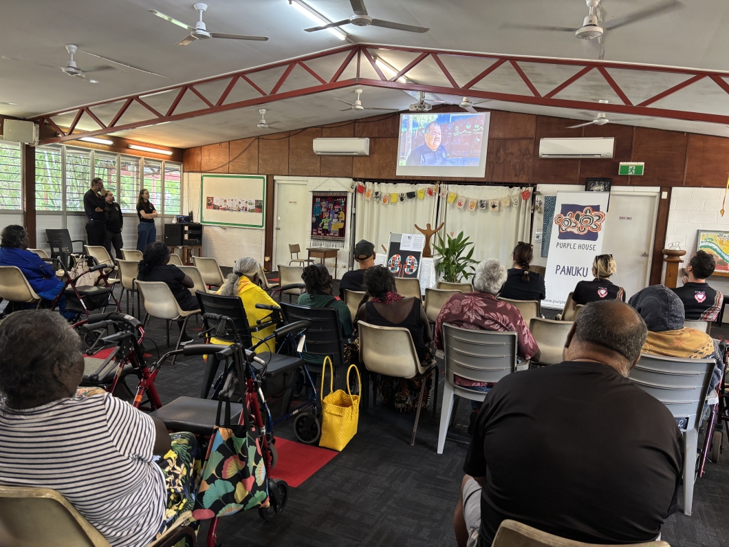 A group of people sit in a community hall, facing a presentation at the front of the room. Banners displayed are for Purple House, and the screen is showing a video of a First Nations man talking about organ and tissue donation.
