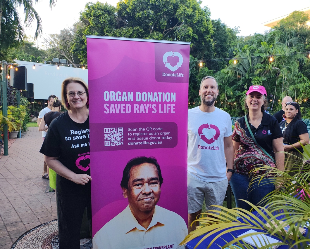 A group of people standing outdoors next to a banner promoting organ donation. The banner reads ‘Organ donation saved Ray's life’ and includes a QR code for registration at donatelife.gov.au. The individuals are wearing shirts and accessories with the DonateLife logo.