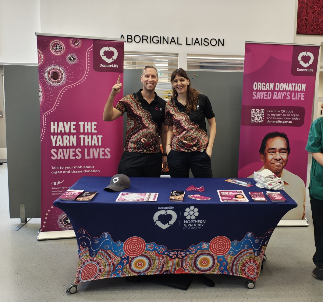2 people stand behind a DonateLife booth promoting organ and tissue donation. They wear matching shirts with Aboriginal designs. The table is covered with a blue cloth featuring the DonateLife and Northern Territory Government logos, along with Aboriginal artwork. Pamphlets and promotional items are displayed. Behind them are 2 banners, one encouraging conversations about donation and another sharing Ray's story with a QR code to register.