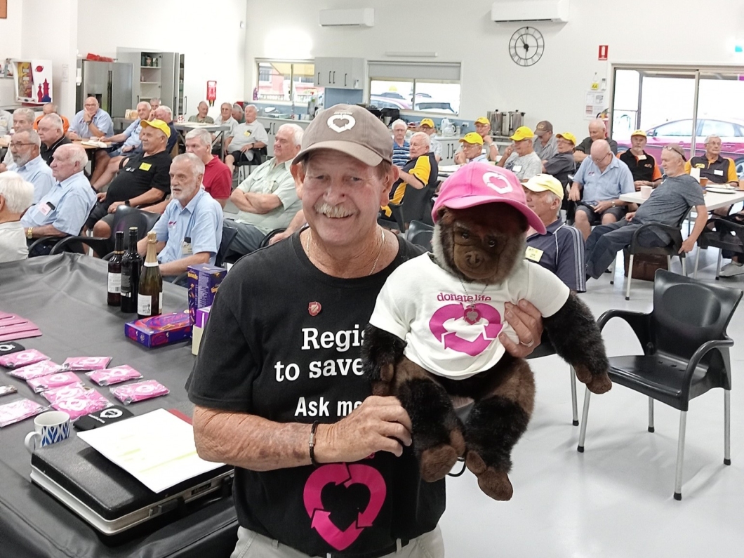 A man wearing a DonateLife branded t-shirt and cap holds a plush monkey dressed in a DonateLife branded t-shirt, surrounded by a group of men seated in a community hall.