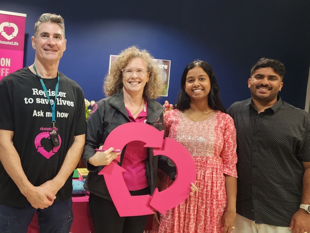Four smiling individuals stand together, holding a large pink heart symbol, promoting organ donation awareness.