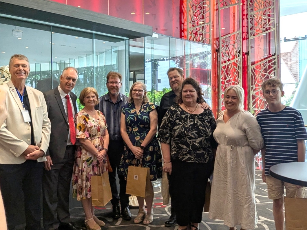 A group of 9 people stand together in a modern lobby, holding gift bags, with a vibrant red and glass backdrop. 
