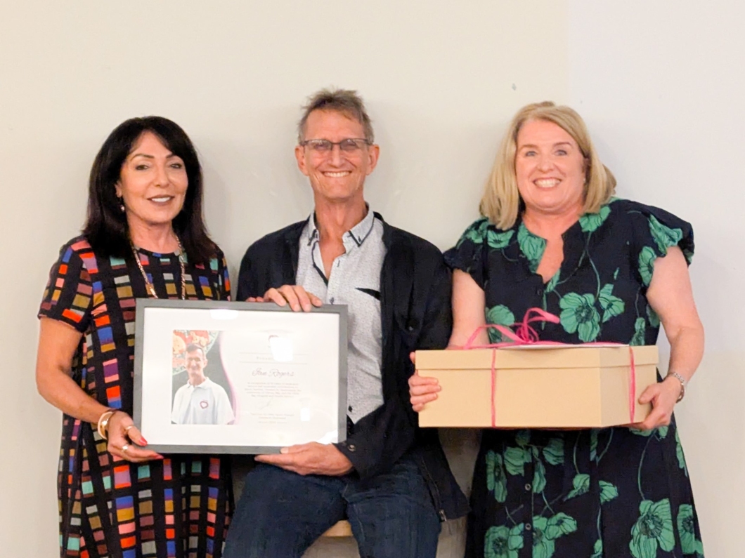 Three individuals pose together, holding a framed certificate and a gift box, celebrating a special occasion.