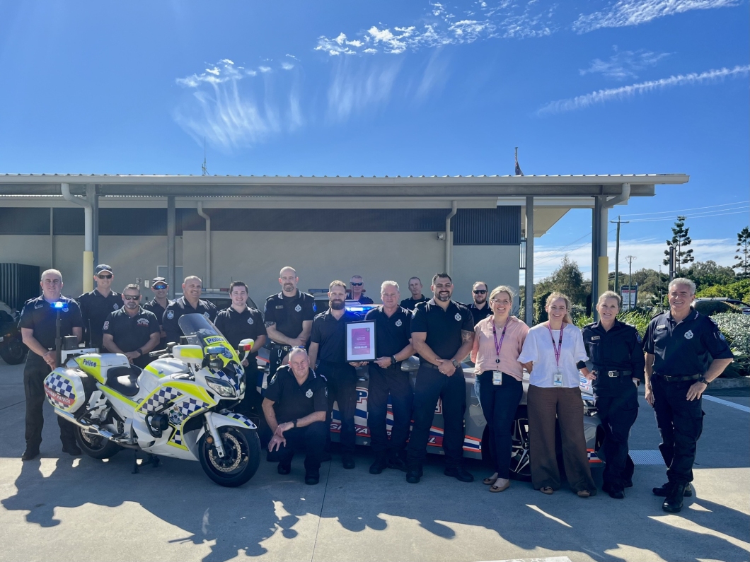A group of police officers and personnel stands together outdoors, alongside a marked police motorcycle and vehicle, under a clear blue sky.