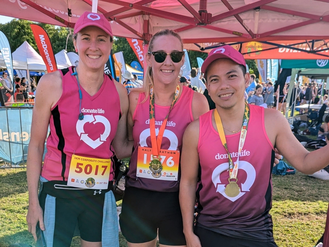 Three runners in magenta DonateLife shirts stand together, proudly displaying their medals and bib numbers at a sports event under a tent.