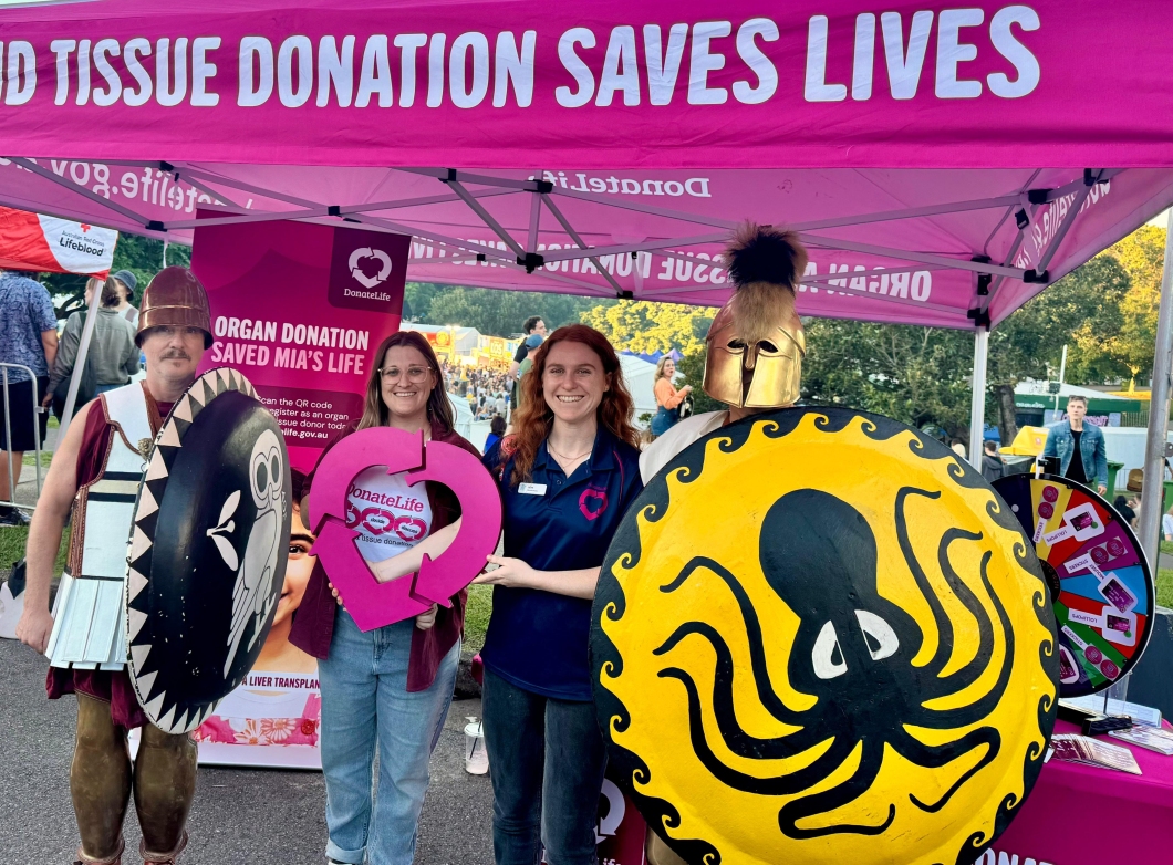A group of people stand under a pink tent promoting organ and tissue donation, holding colourful shields and a heart-shaped sign.