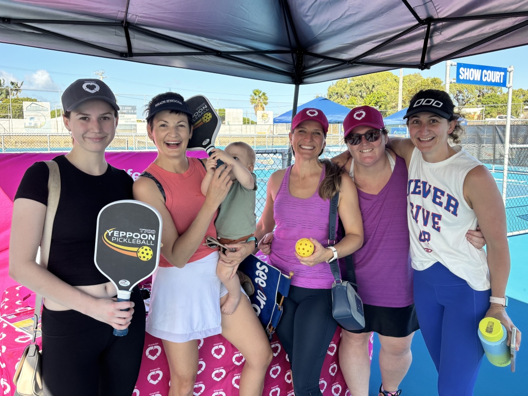 A group of 5 women and a young child stand under a canopy, holding pickleball paddles and balls, smiling with a colourful background of tennis courts.