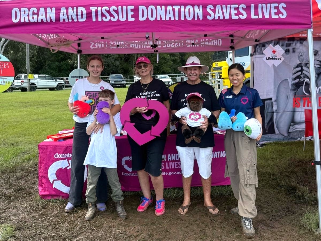  Five individuals pose under a pink tent promoting organ and tissue donation, holding colorful plush toys and smiling at the camera.