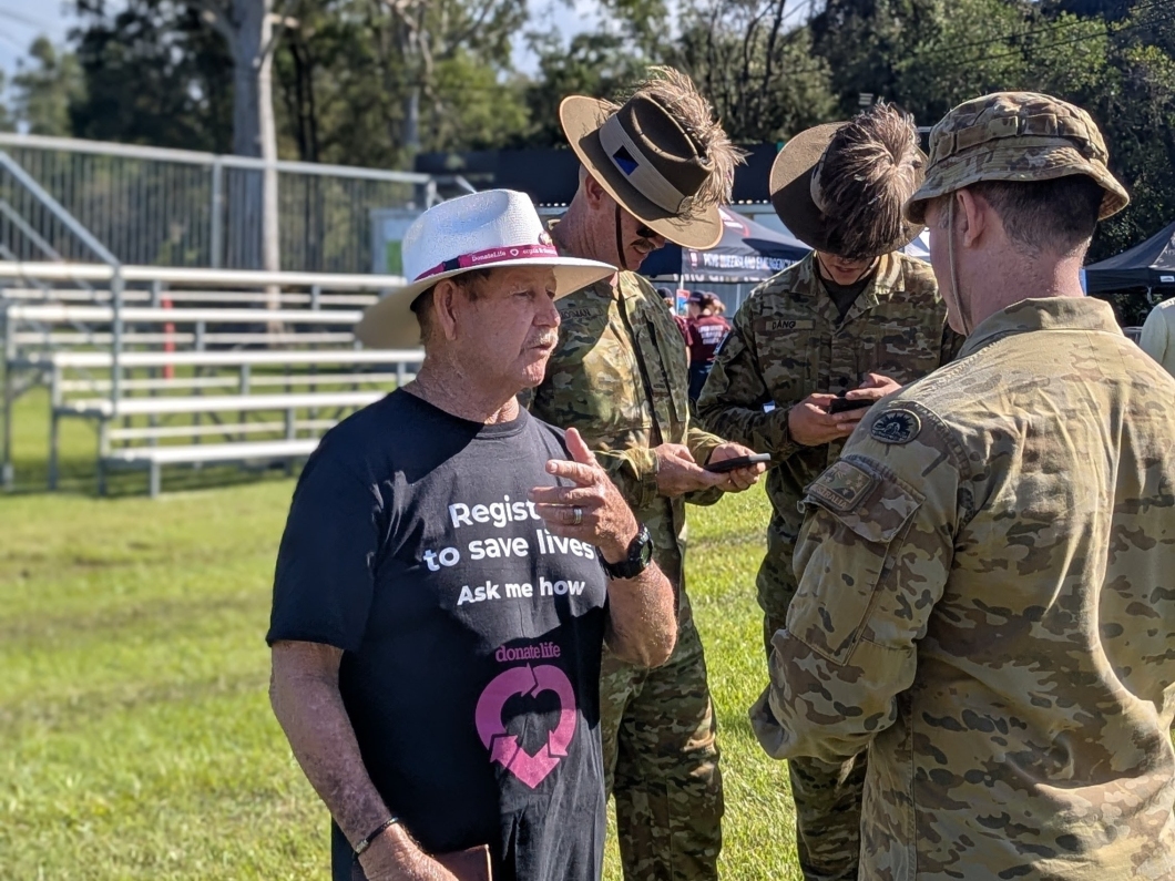 A man in a black DonateLife t-shirt speaks with a man in an army uniform, while 2 more military personnel stand behind them, using their phones to register as organ and tissue donors.