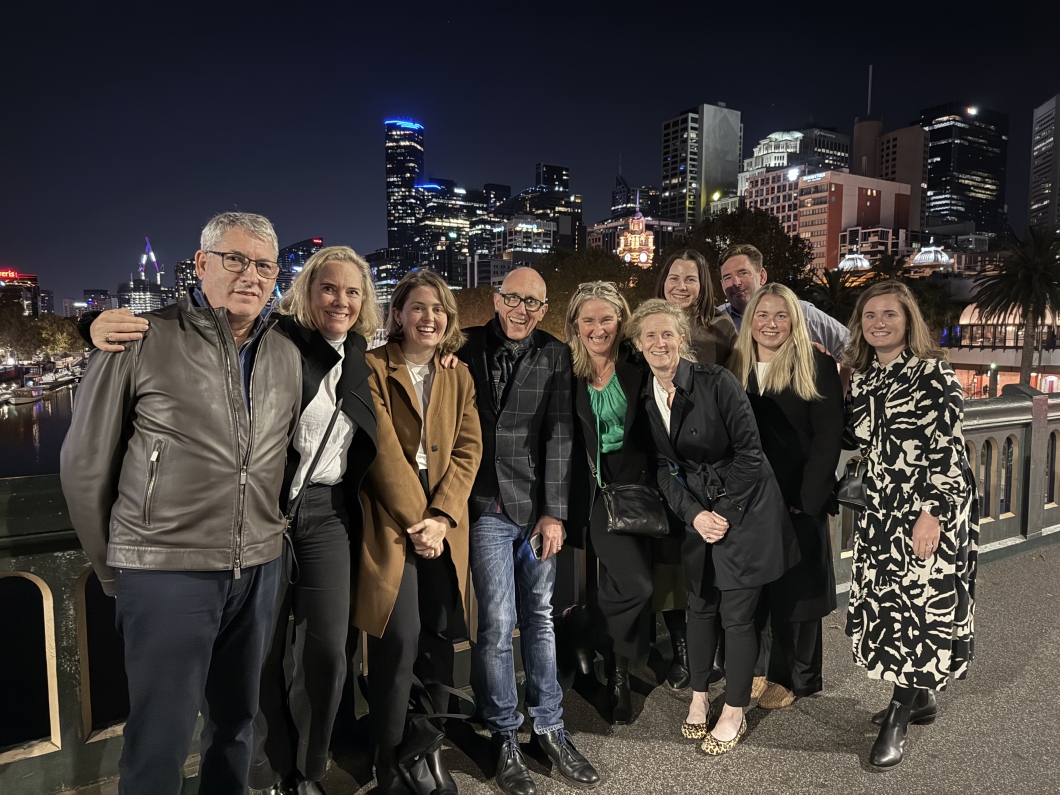 A group of 10 people standing together on a bridge at night, with a city skyline and lights in the background.