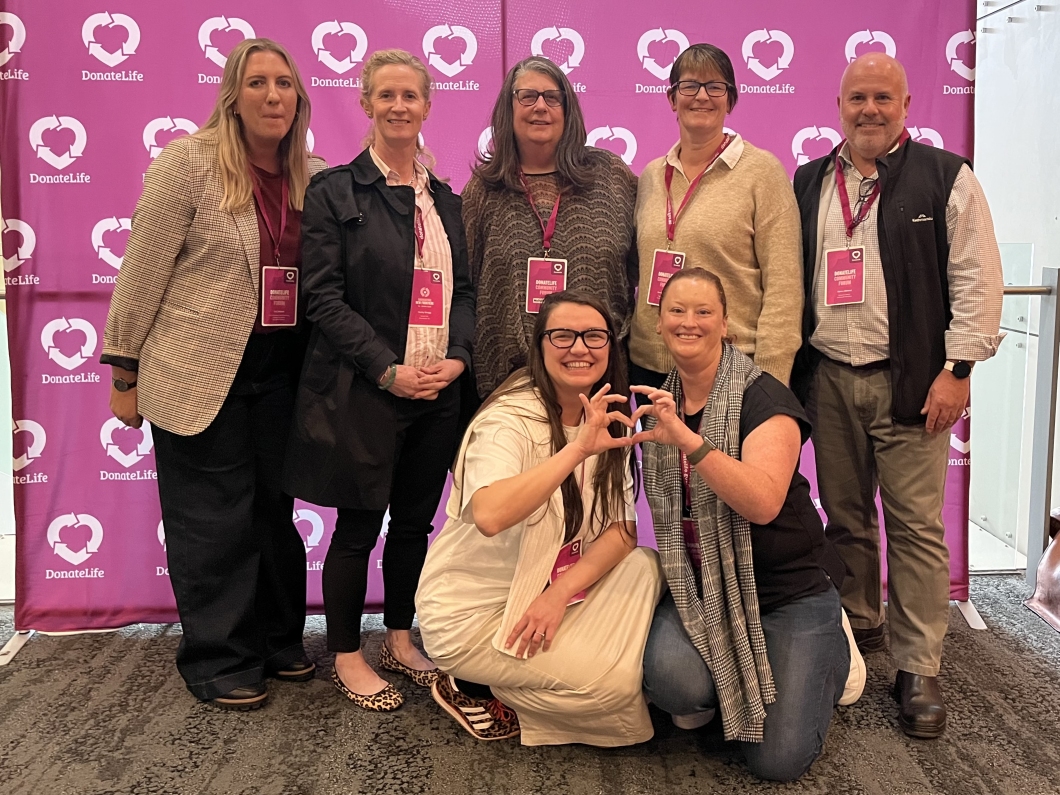A group of 7 people stand in front of a large magenta DonateLife background banner at an event, all wearing name tags and smiling.