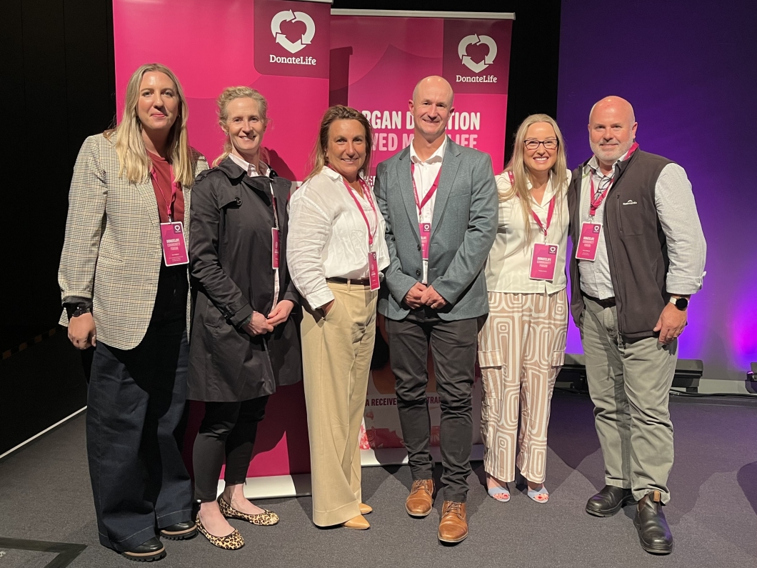 A group of 7 people stand in front of pink DonateLife banners at an event, all wearing name tags and smiling.