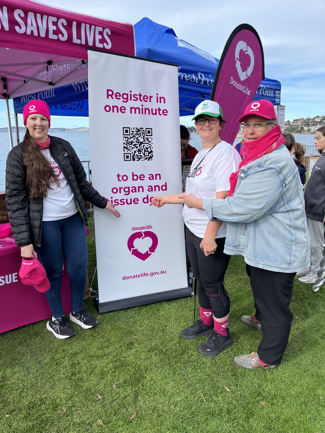 A group of people at an outdoor event promoting organ and tissue donation, with a pink and white banner that reads 'Register in one minute to be an organ and tissue donor.' The banner includes a QR code, the DonateLife logo, and the website donatelife.gov.au. Tents and additional branded banners are visible in the background.