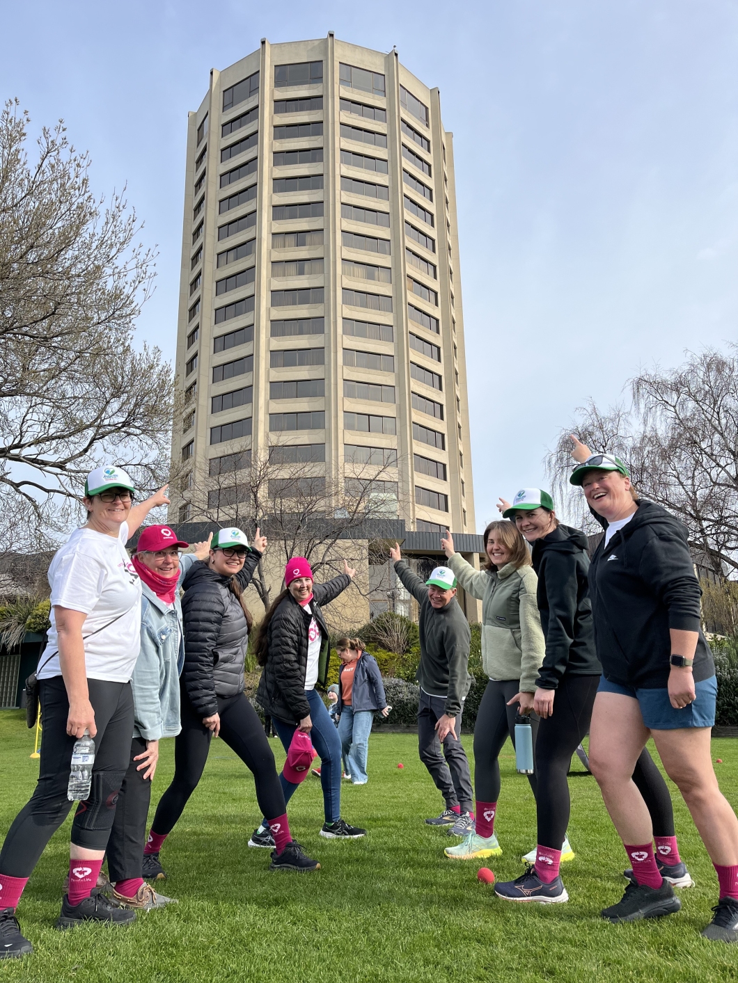 A group of people stand outdoors in front of a tall, multi-story building with vertical columns and many windows. Some individuals are pointing upward toward the building. Trees frame the scene on both sides.