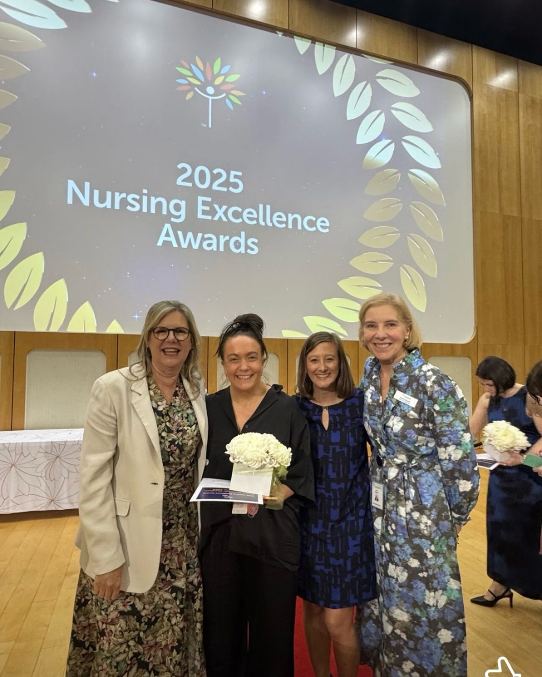 Four women pose together at the 2025 Nursing Excellence Awards, holding bouquets and smiling in front of a decorated presentation backdrop.