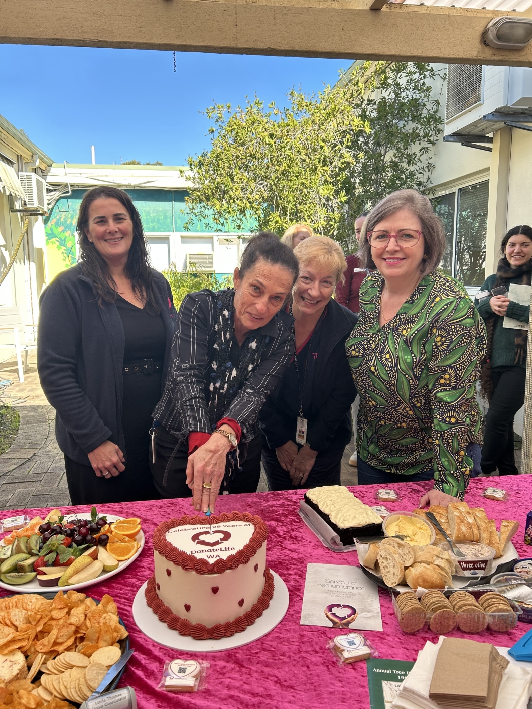 4 people stand in front of a table covered with plates of food. The person in the middle is cutting into a red and white cake in the shape of a heart. In the background, other people are visible, as well as buildings and trees.