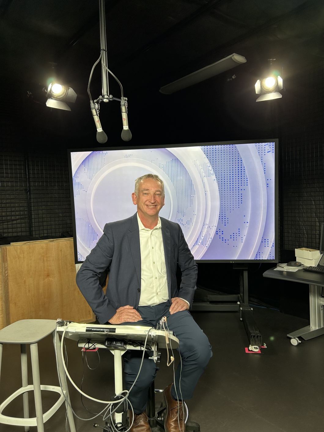 A person sits in a studio setting with professional lighting and ceiling-mounted microphones. Behind them is a large screen displaying circular and dotted graphic designs, suggesting a news or broadcast environment.