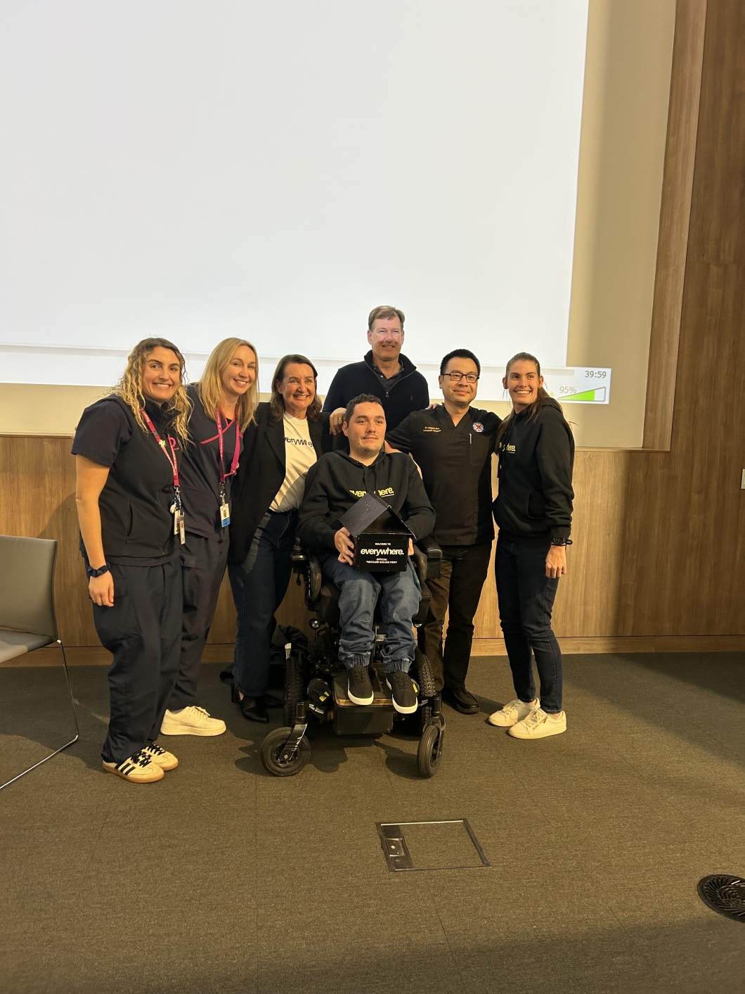 7 people stand together in front of a large projection screen. One person is seated in a motorised wheelchair holding a box labeled 'everywhere.' 