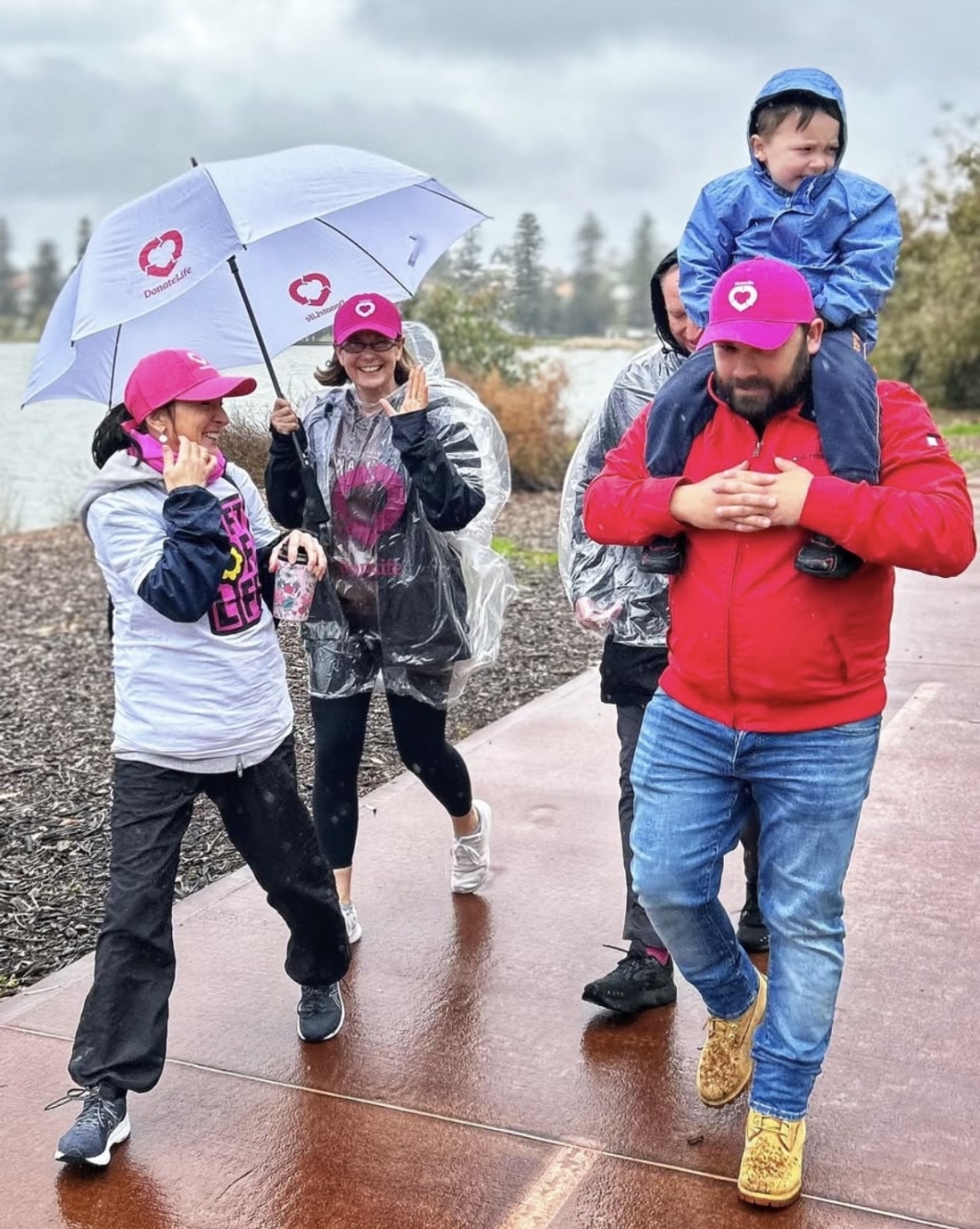 A group of people walk along a wet pathway on a rainy day, wearing rain ponchos and holding umbrellas with the 'DonateLife' logo. One person carries a child on their shoulders.  