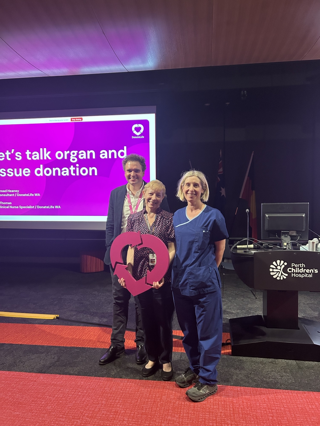 3 individuals stand in front of a presentation screen displaying the title 'Let's talk organ and tissue donation' with names and titles of speakers from DonateLife WA. They hold a large pink DonateLife logo. A podium with the Perth Children's Hospital logo is also visible.