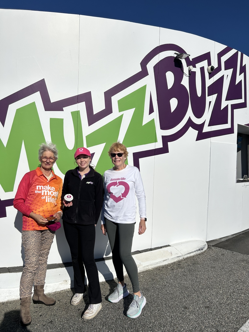 3 people stand in front of a building with a large 'Muzz Buzz' sign in green and purple. One wears an orange shirt with the text 'make the most of life!', another wears a black jacket and pink hat holding a takeaway coffee cup, and the third wears a white DonateLife shirt.