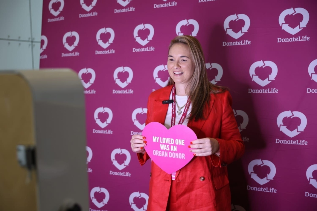 A person in a red blazer holds a heart-shaped sign that says, ‘My loved one was an organ donor’, against a DonateLife backdrop.