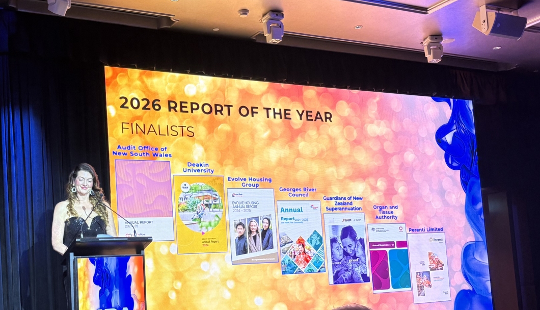 A presenter stands at a lectern on a stage beside a large illuminated screen displaying ‘2026 Report of the Year Finalists’. The screen shows several annual report covers from organisations including the Audit Office of New South Wales, Deakin University, Evolve Housing Group, Georges River Council, Guardians of New Zealand Superannuation, Organ and Tissue Authority, and Perenti Limited, against a bright orange and pink abstract background. Overhead lighting and speakers are visible above the stage.