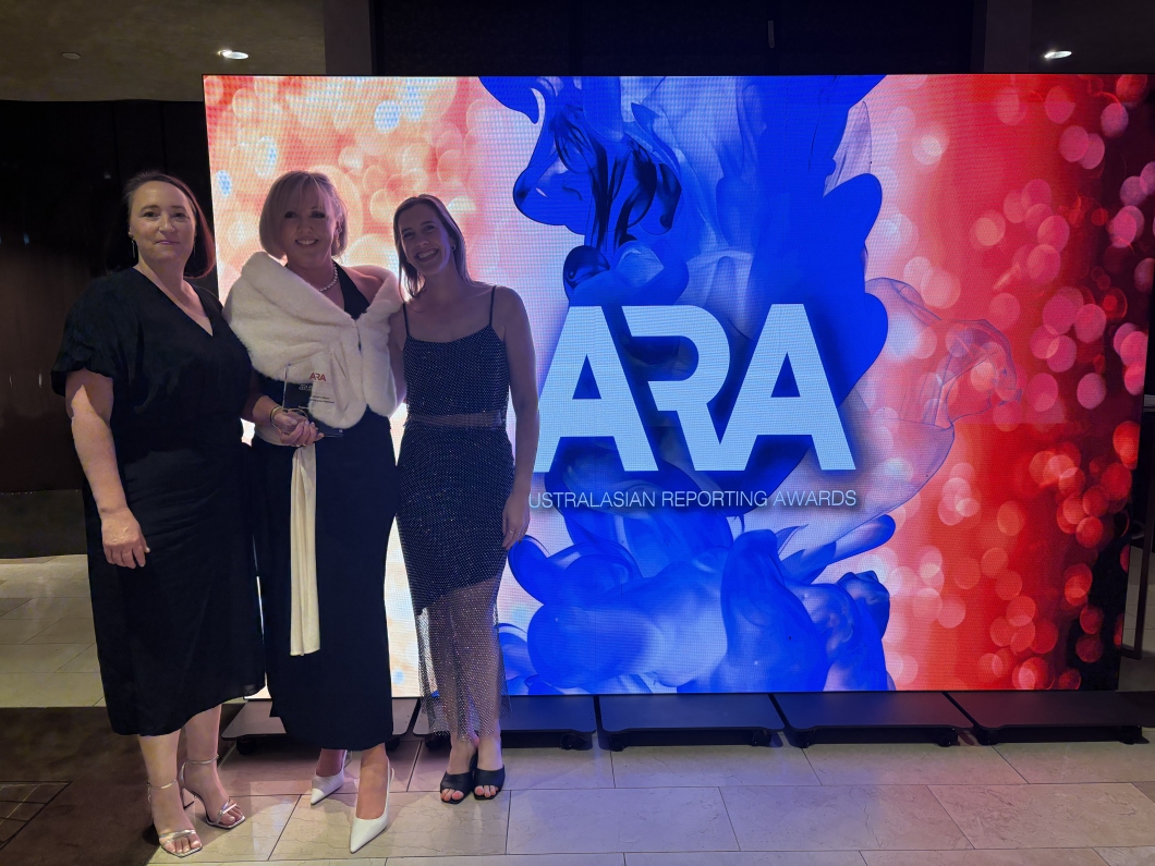 3 people pose together in front of a large illuminated backdrop displaying ‘ARA Australasian Reporting Awards’. One person holds a glass award while wearing a dark outfit with a light shawl, and the other 2 stand on either side in formal attire. The colourful background features blue and red abstract graphics and bright lighting, with the group standing on a tiled floor at an event venue.