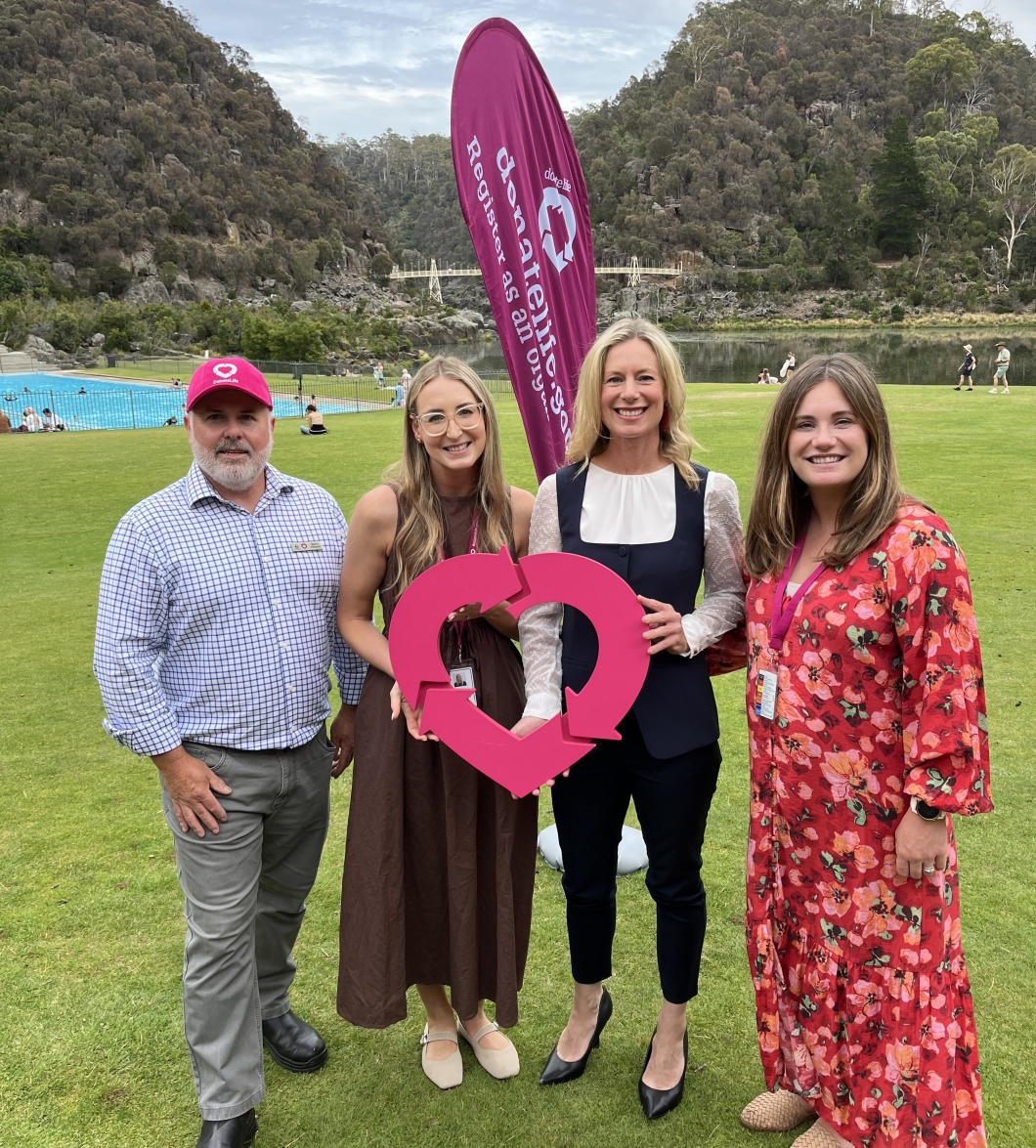 4 people stand together on a grassy field holding a large DonateLife wooden logo. Behind them is a tall purple DonateLife banner and a scenic backdrop of hills, trees, and a body of water.