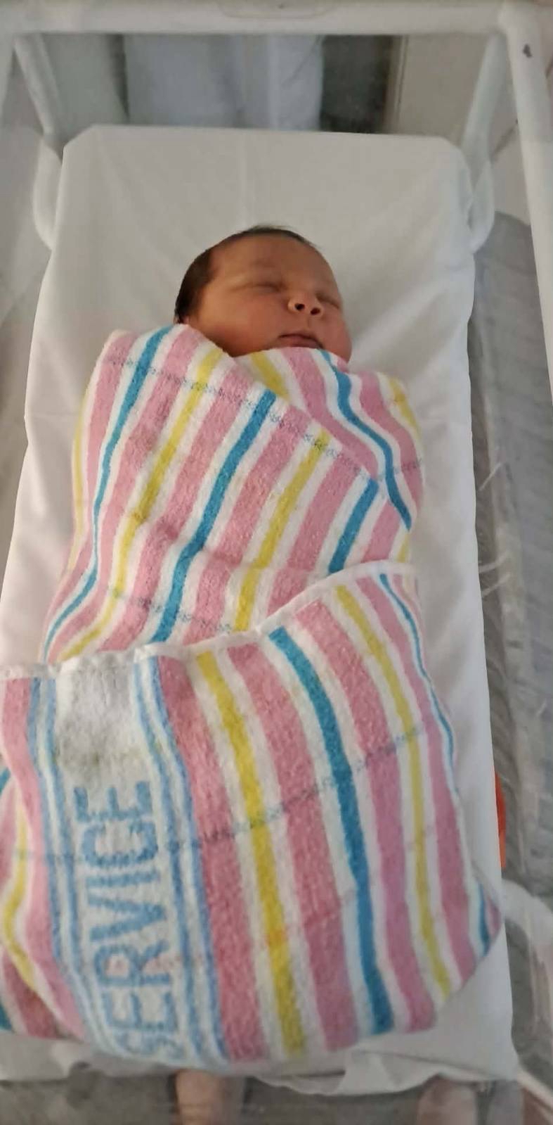 A baby swaddled in a colourful striped blanket sleeps in a hospital crib.