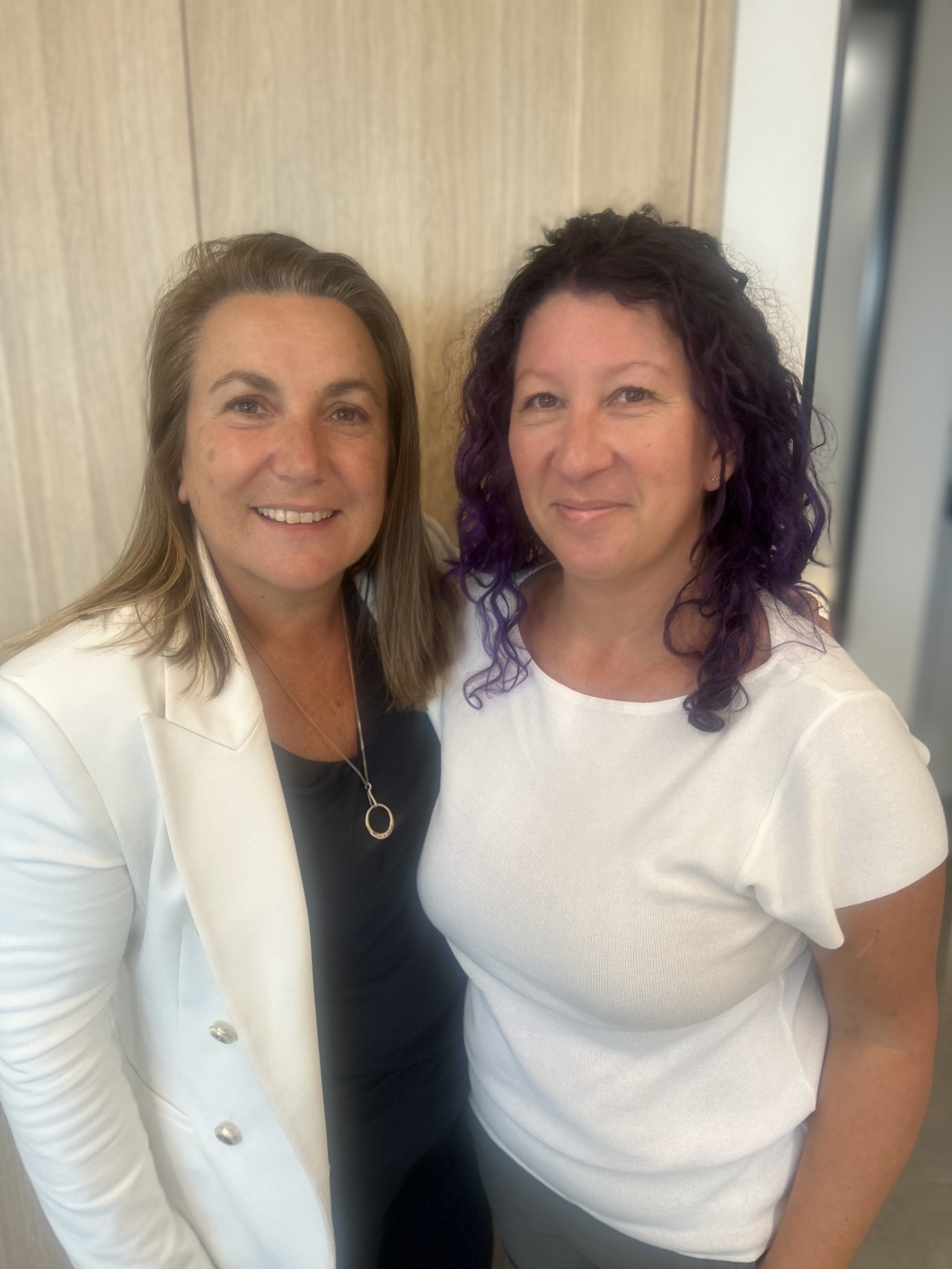 2 women, one in a white blazer and black top and the other in a white top, stand close together and smile at the camera. 