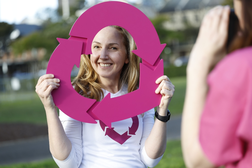 A person wearing a white DonateLife shirt holds a large magenta DonateLife logo frame in front of their face while posing outdoors. Another person is visible in the foreground taking a photo. The background shows a blurred park setting with grass, trees and buildings. Service NSW supported the campaign through registration prompts in the