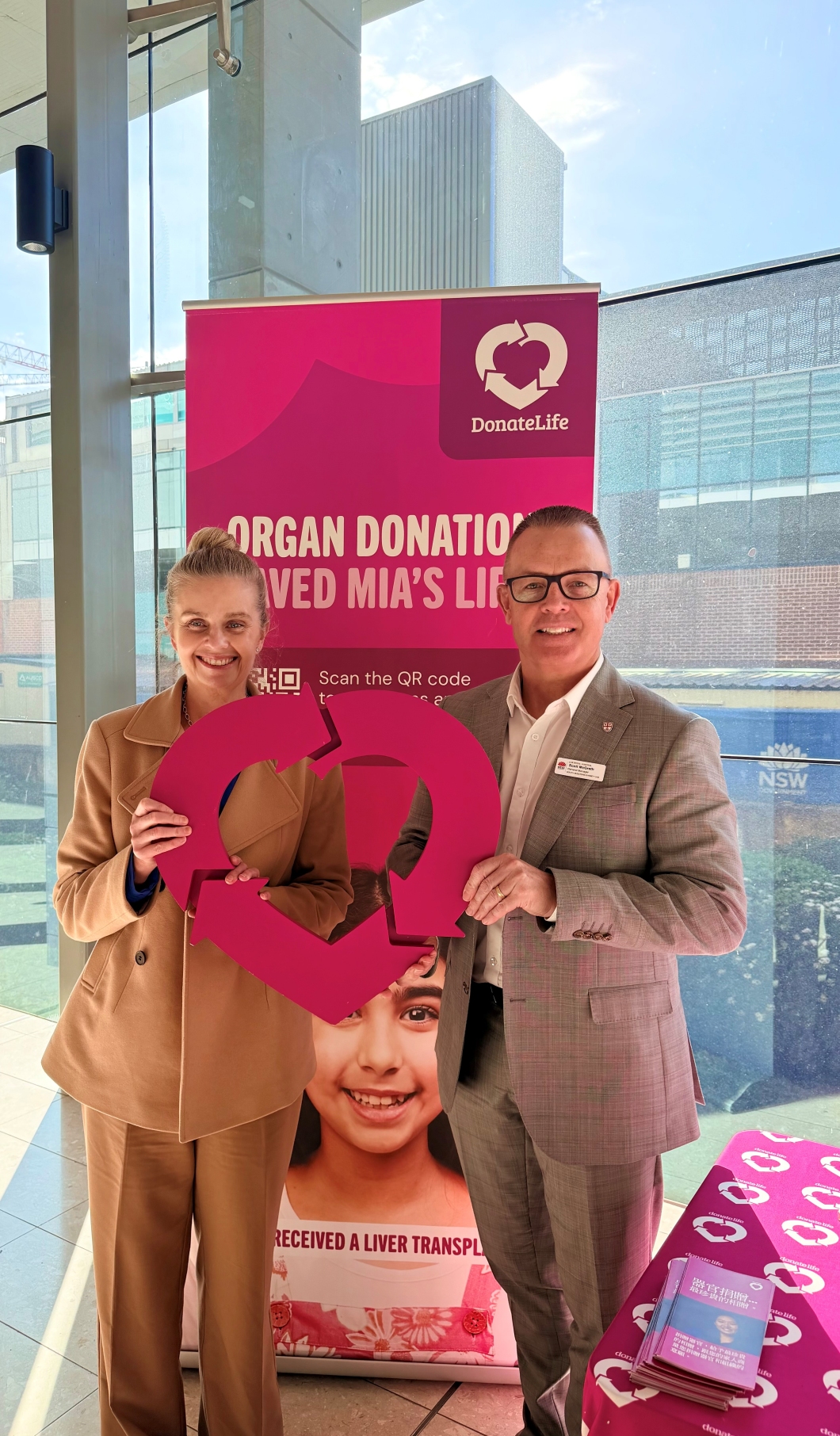2 people stand in front of a DonateLife display at a hospital, holding a large magenta DonateLife heart-shaped logo. Behind them, a banner reads ‘Organ donation saved Mia’s life’ and includes a QR code and photo of a transplant recipient. A table covered with DonateLife-branded materials is visible beside the display, with large windows and hospital buildings in the background.