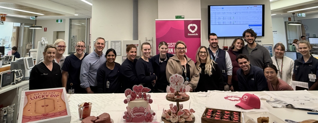 A group of hospital staff pose behind a table decorated for a DonateLife bake-off competition in a hospital ward. The table displays magenta-themed cakes, cupcakes, brownies and DonateLife promotional items, including a magenta cap and a ‘Gift of Life Lucky Dip’ box. A DonateLife banner stands behind the group, while medical equipment, patient care areas and digital screens are visible throughout the clinical setting.