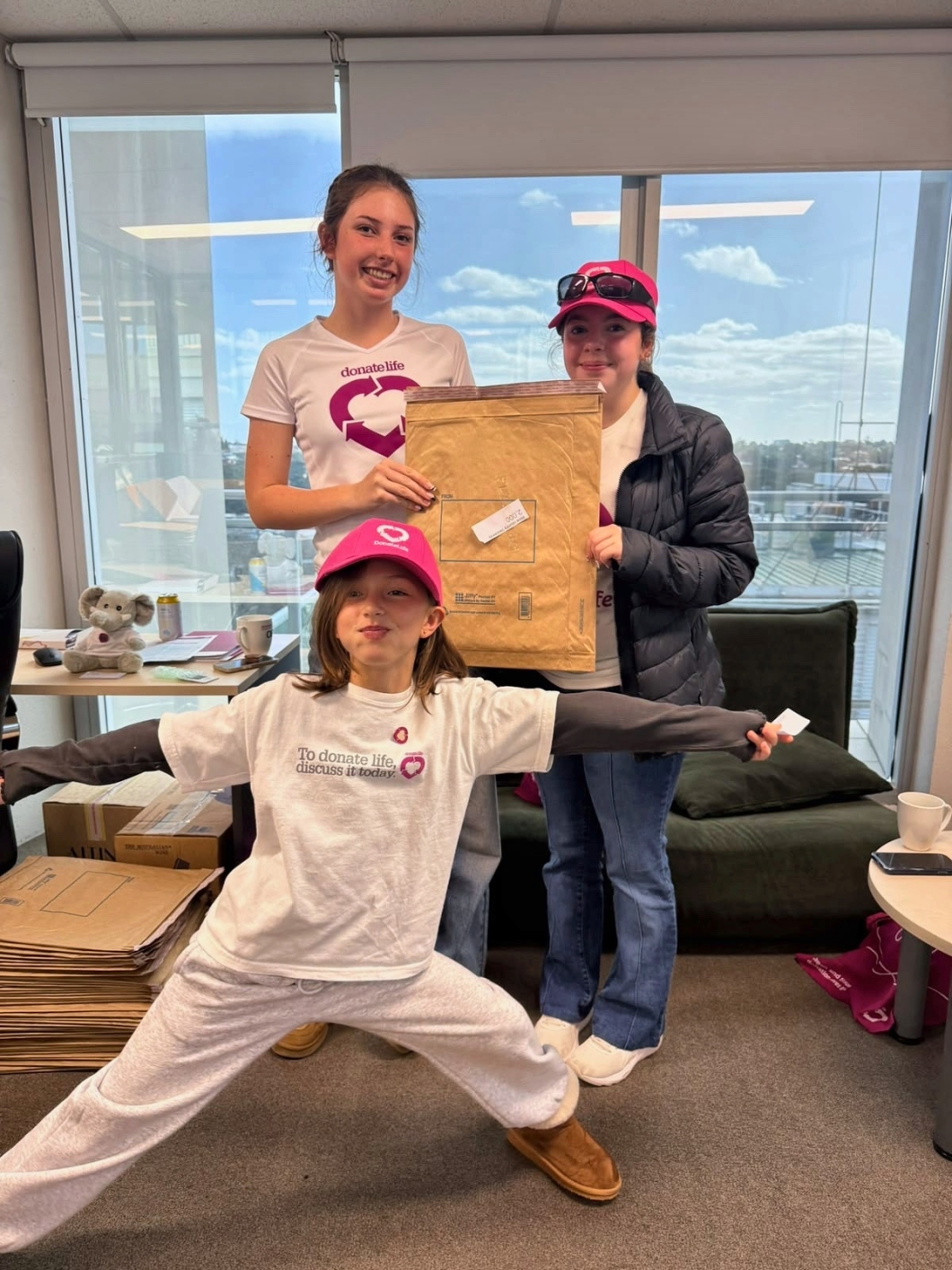 3 young DonateLife volunteers stand in an office packing information materials into large padded envelopes. One volunteer in a DonateLife T-shirt and pink cap poses with arms outstretched in the foreground, while 2 others stand behind holding a large mailing envelope. Stacks of flat-packed envelopes and office furniture are visible around the room, with large windows showing a cloudy blue sky in the background.