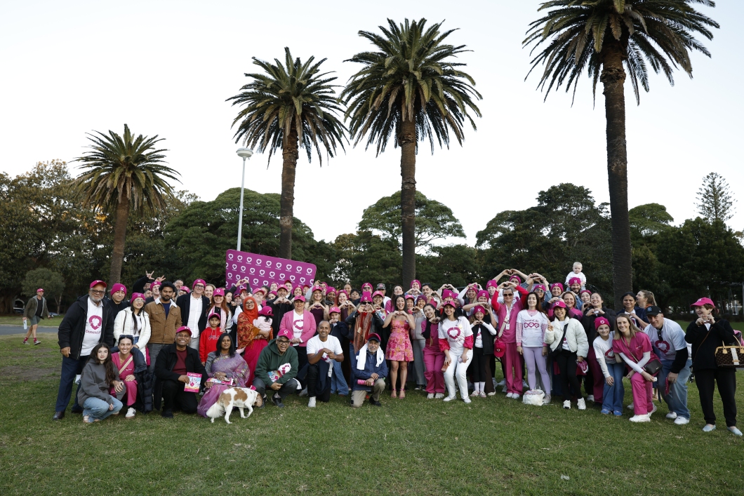  A large group of DonateLife supporters gather in a park for a community event, standing and kneeling on the grass beneath tall palm trees. Many participants wear magenta DonateLife hats, shirts and accessories, and several people form heart shapes with their hands. A magenta DonateLife media wall is visible behind the group, along with a dog in the front row and parkland in the background.