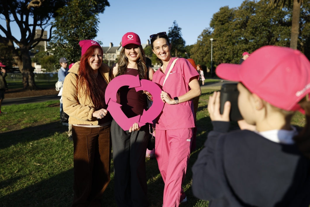 3 DonateLife supporters pose in a park while holding a large magenta DonateLife heart-shaped logo. Two wear magenta DonateLife hats and one is dressed in pink scrubs. In the foreground, a child in a magenta cap takes their photo. Trees, grass and other event participants are visible in the background at an outdoor DonateLife community event.