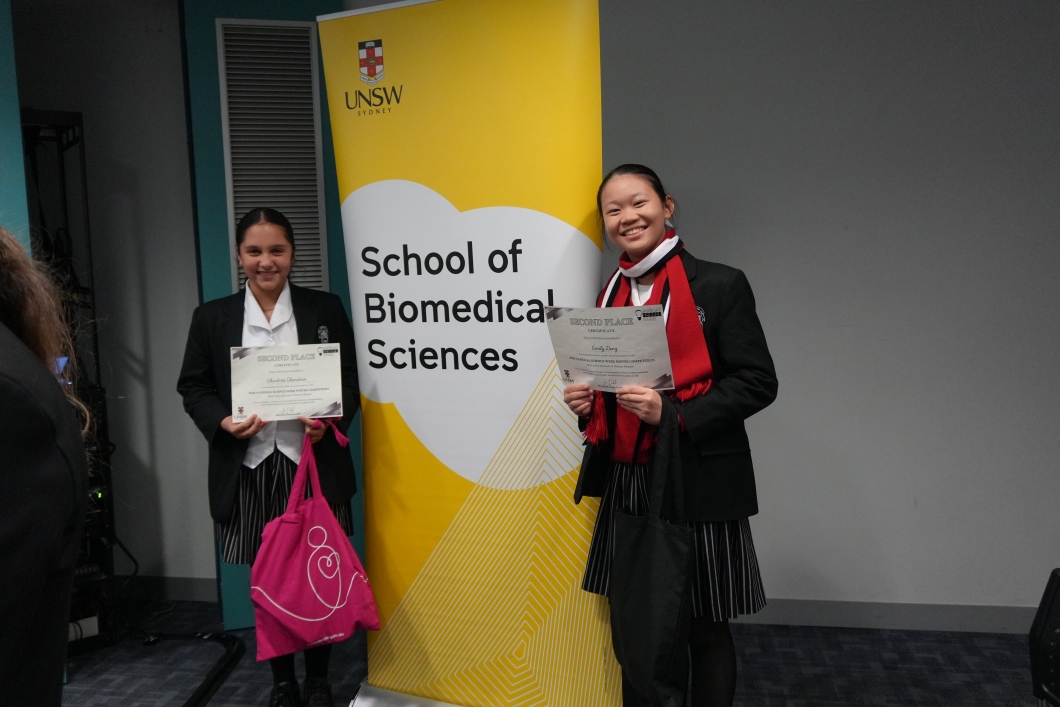 2 students stand in front of a University of New South Wales (UNSW) School of Biomedical Sciences banner, holding certificates for second place in a science competition. One student also holds a bright magenta DonateLife tote bag. The students are wearing school uniforms and posing in an indoor presentation space with the UNSW banner prominently displayed behind them.