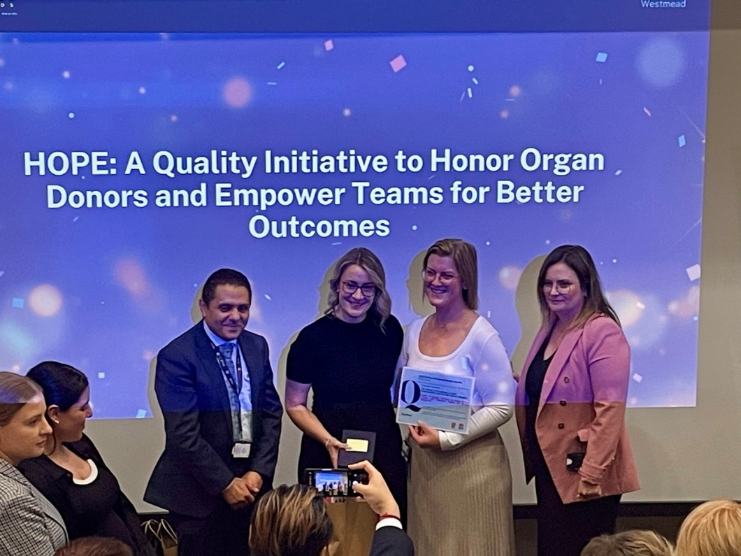 A group of people stand in front of a screen displaying the message: 'HOPE: A Quality Initiative to Honor Organ Donors and Empower Teams for Better Outcomes.' One person holds a certificate or award, while another takes a photo with a smartphone. The background features a blue gradient with light spots and confetti-like elements.