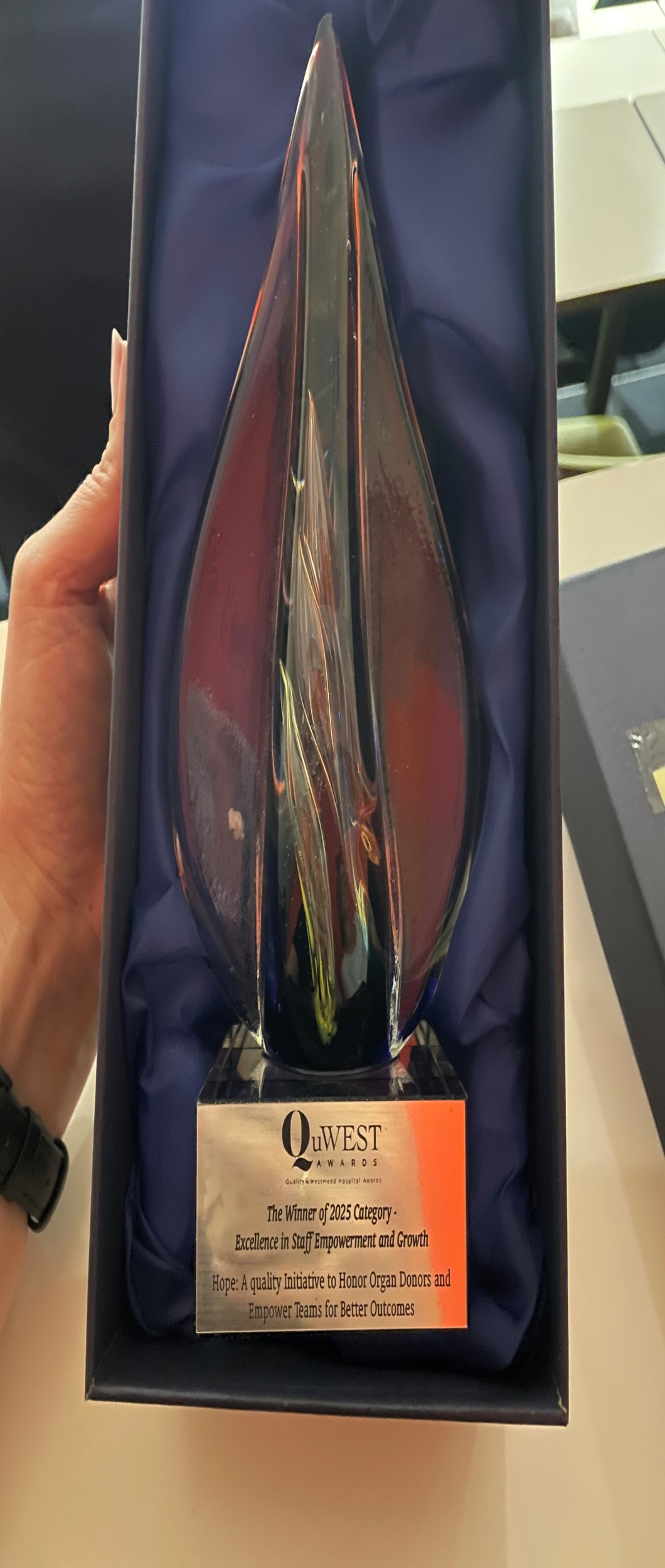 A hand holds a trophy in a box lined with blue fabric. The glass trophy has an abstract flame-like design in red and blue hues. The engraved plaque reads: 'QuWEST AWARDS – The Winner of 2025 Category – Excellence in Staff Empowerment and Growth – Hope: A quality initiative to Honor Organ Donors and Empower Teams for Better Outcomes.'
