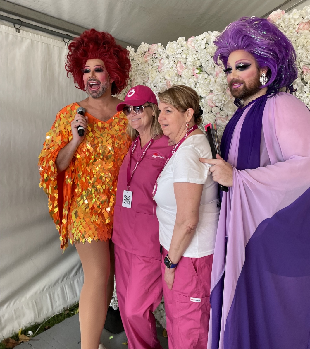 2 drag queens in colorful outfits and large wigs stand on either side of 2 people wearing pink uniforms and lanyards, posing together under a white marquee tent. A decorative wall of white flowers forms the backdrop.