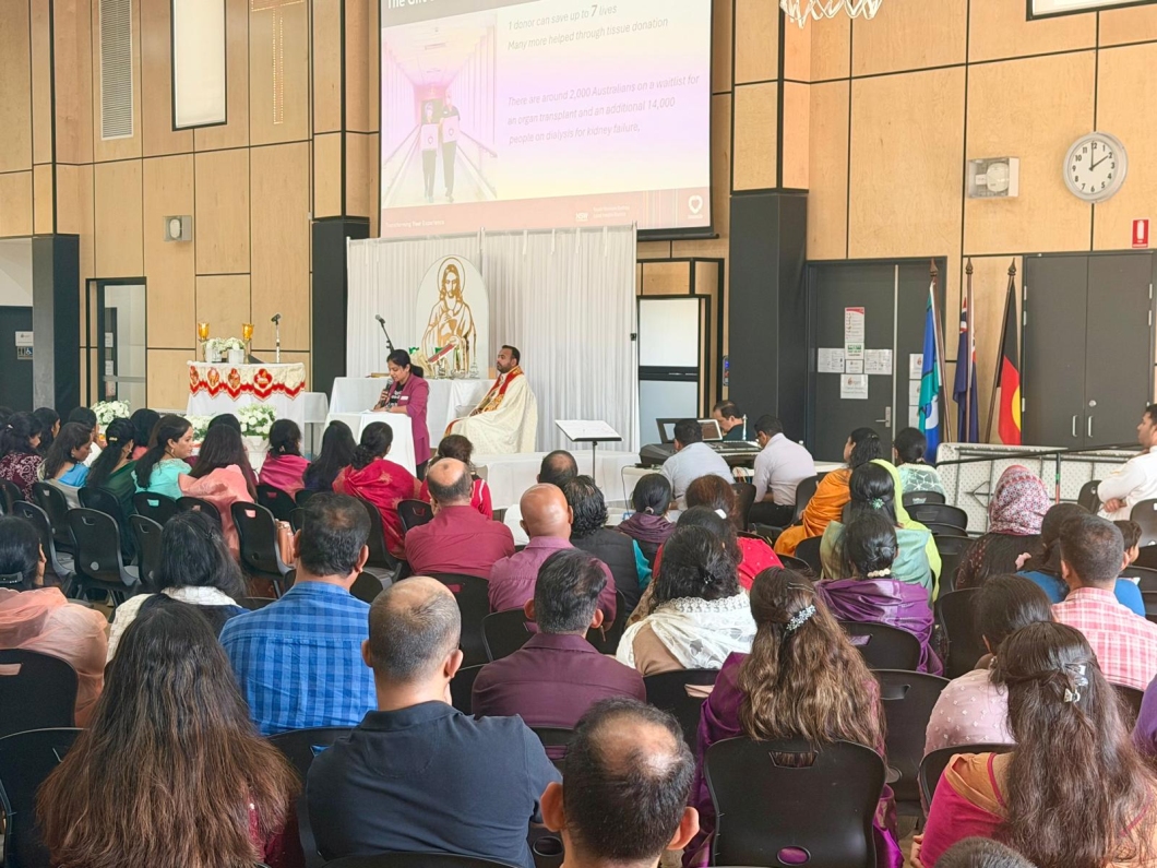 Large community gathering in a hall where rows of seated attendees face a stage. At the front, a speaker stands at a lectern beside a person in ceremonial attire, with an altar-like setup and microphones nearby. Behind them, a large projection screen displays information about organ donation, including text about transplant waitlists. Flags stand to one side of the stage, and a wall clock and wood-paneled walls are visible in the background.