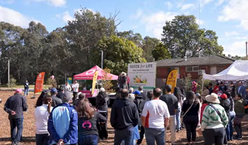 Group of people gathered outdoors at an event with tents and banners, including one that reads ‘SEEDS for LIFE’, set in a park-like area with trees in the background.