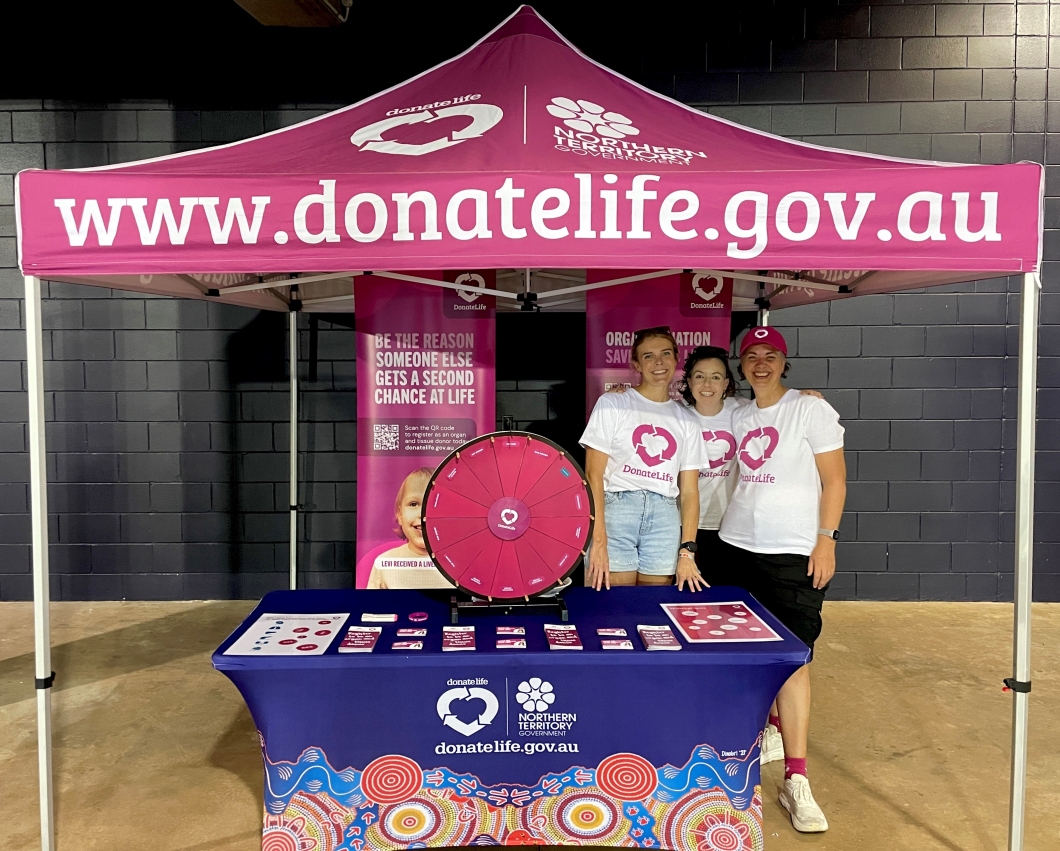 3 people stand behind a DonateLife information stall under a magenta canopy printed with ‘www.donatelife.gov.au’ and Northern Territory Government logos. The table displays brochures, registration materials, and a magenta spinning prize wheel. Behind them are vertical banners with messages about organ donation, including ‘Be the reason someone else gets a second chance at life’.