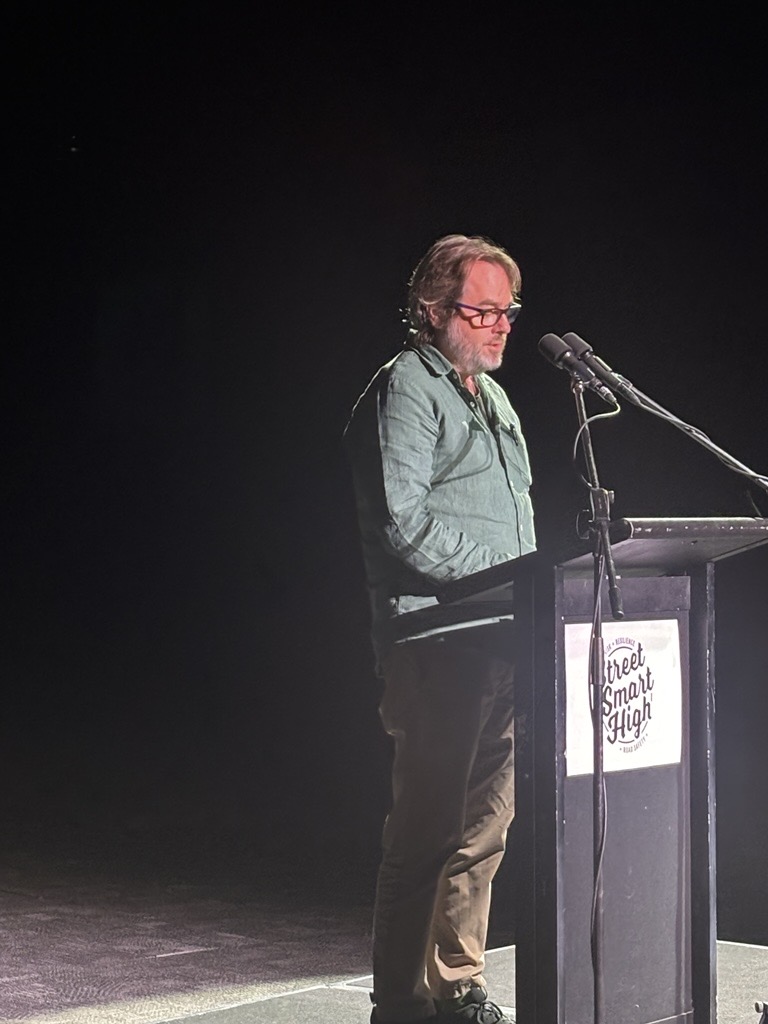 A person stands at a lectern on a dimly lit stage, speaking into 2 microphones. The lectern displays a ‘Street Smart High’ sign, and stage lighting highlights the speaker against a dark background.