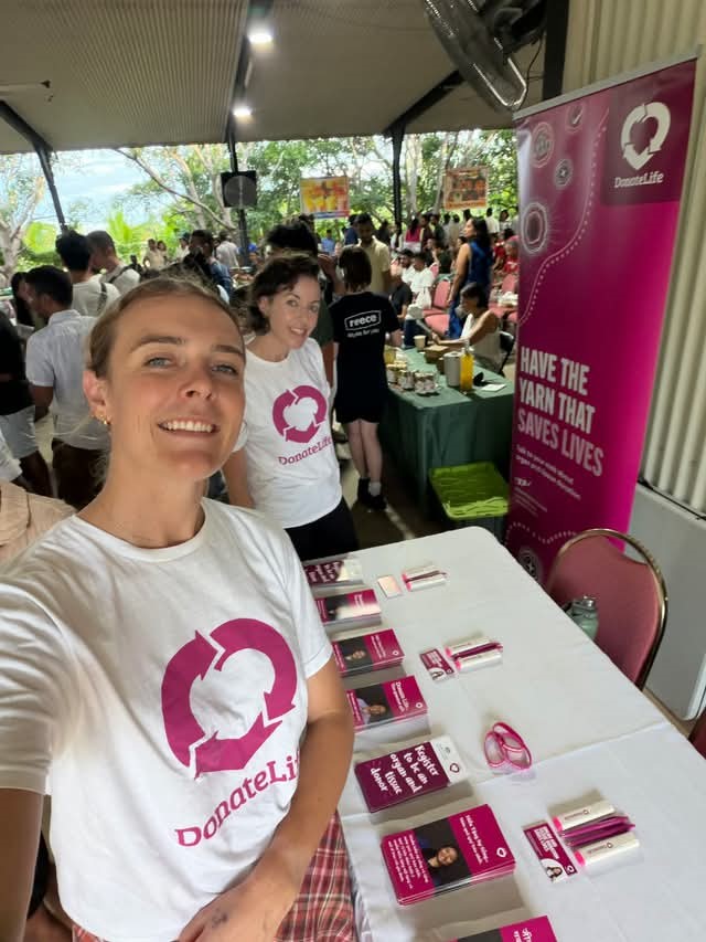 2 people wearing DonateLife shirts stand behind an information table at a busy indoor event, taking a selfie. The table is covered with brochures, pens, and registration materials. A tall pink banner reads ‘Have the yarn that saves lives’ alongside the DonateLife logo. In the background, a crowd of attendees gathers around food stalls and seating areas under a covered open-air space.