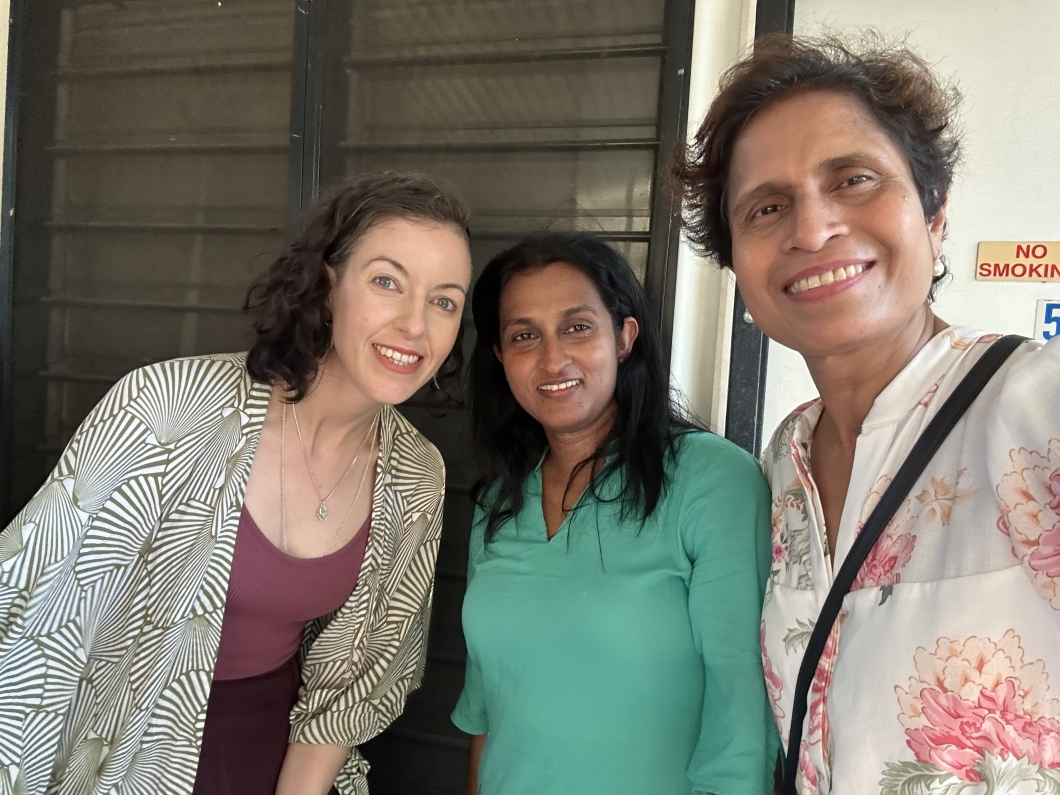 3 people pose closely together for a selfie indoors near a window with metal bars. The individuals are dressed casually, and the person in front holds the camera while the others lean in beside them.