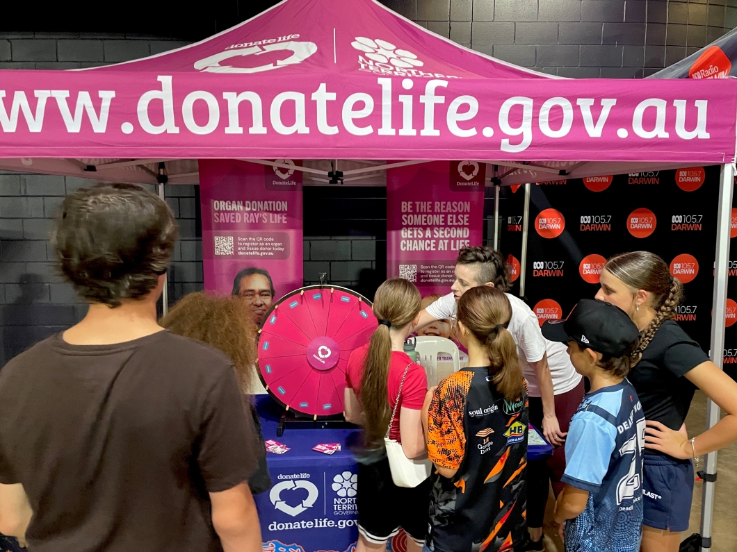A crowd gathers around a DonateLife information stall under a magenta canopy printed with ‘www.donatelife.gov.au’. Staff members engage with attendees near a table displaying brochures and a magenta spinning prize wheel. Behind the stall are banners promoting organ donation, including messages such as ‘Be the reason someone else gets a second chance at life’, and a backdrop featuring ‘ABC 105.7 Darwin’ logos.