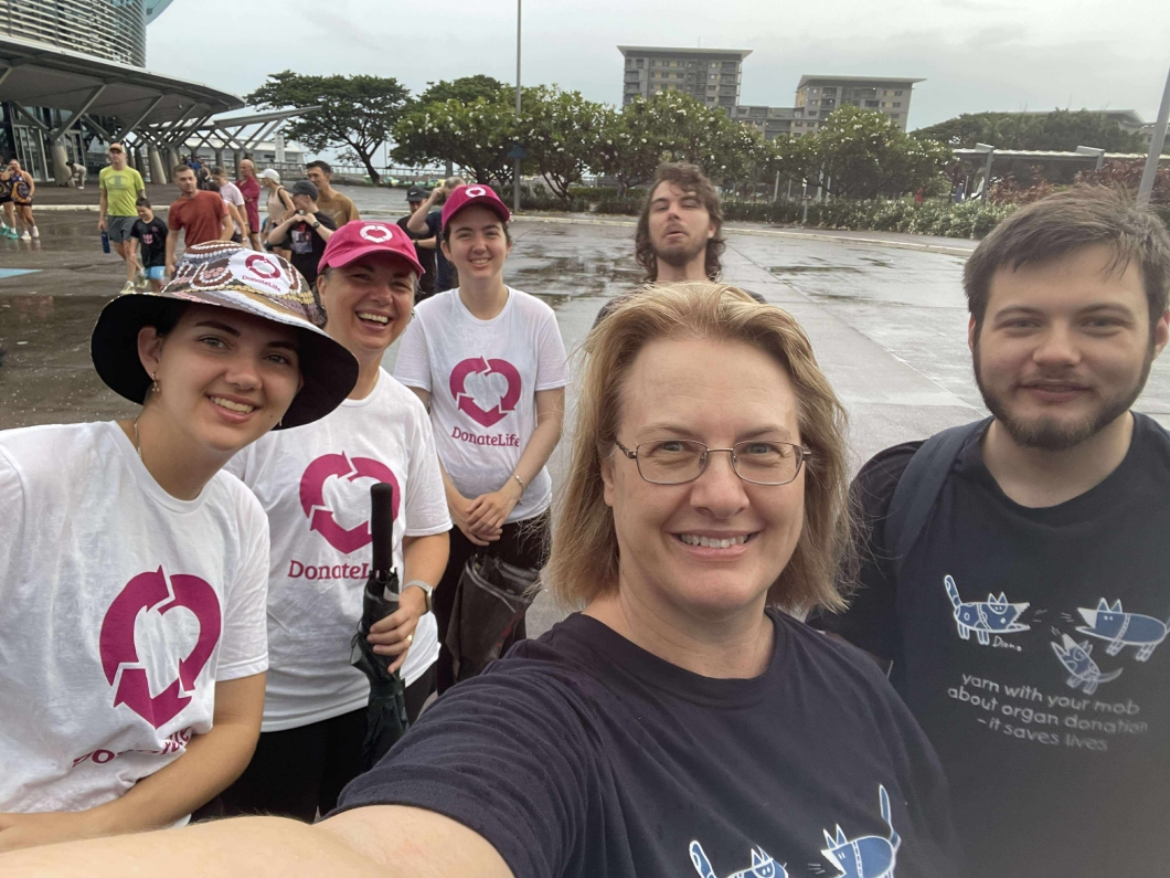 A group of people wearing DonateLife shirts and hats gather outdoors on a wet pavement, posing for a group photo. Some are holding umbrellas. Behind them are trees, parked cars, and tall buildings in the distance, with other event participants walking through the area.