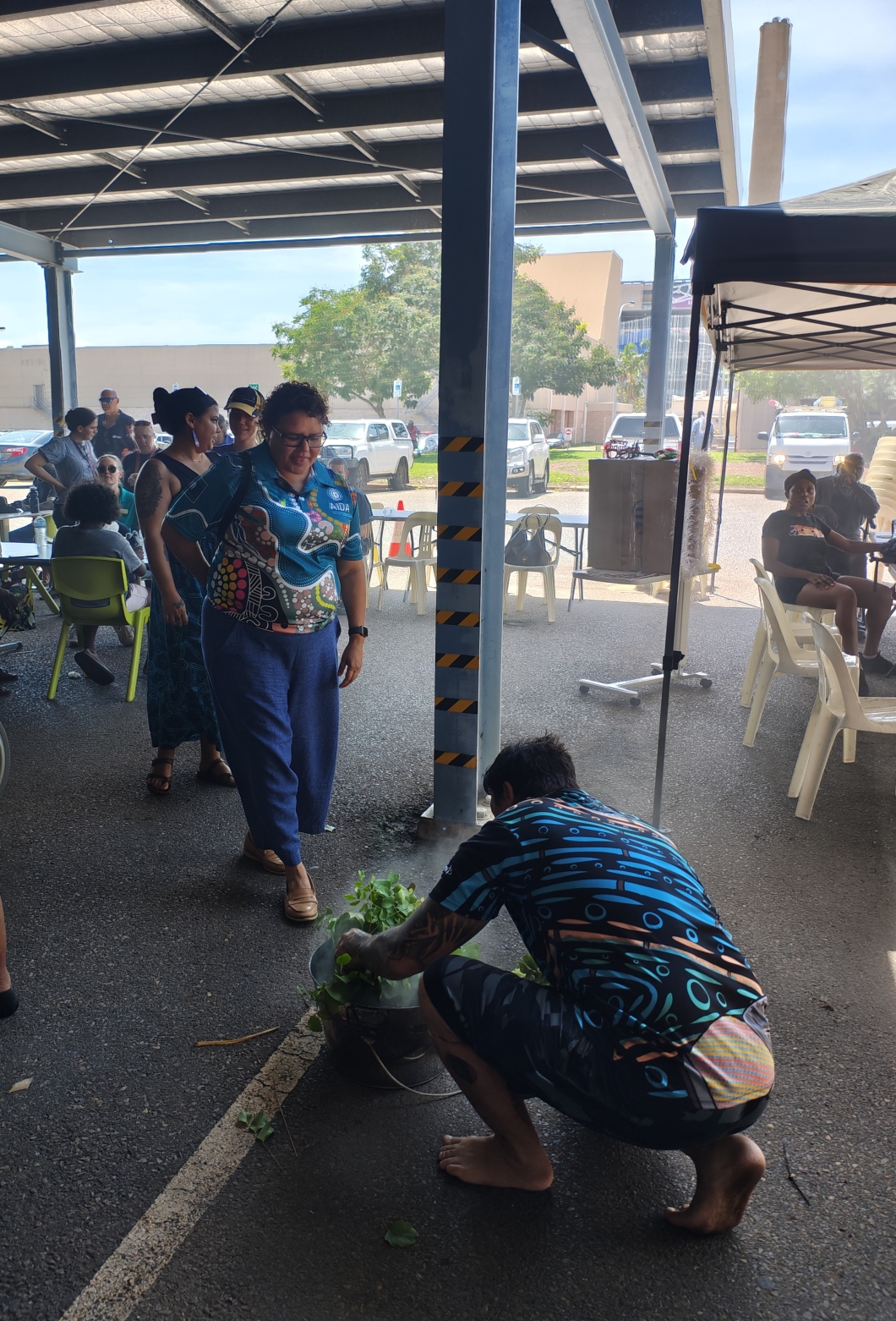 People gather under a covered outdoor area where an event is taking place. Several individuals are seated at tables in the background. In the foreground, one person is crouching down and placing green leafy branches into a smoking metal pot on the ground, creating a cleansing or ceremonial effect. Another person stands nearby watching. The scene is shaded, with parked cars and trees visible in the background beyond the shelter.